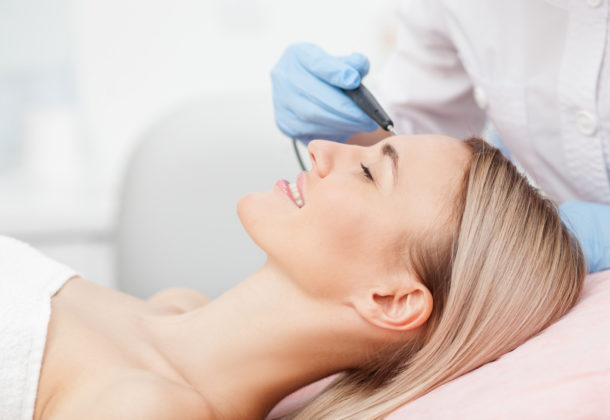 Close up of arms of experienced beautician undergoing laser skin treatment of skin on female face. The young woman is lying and smiling